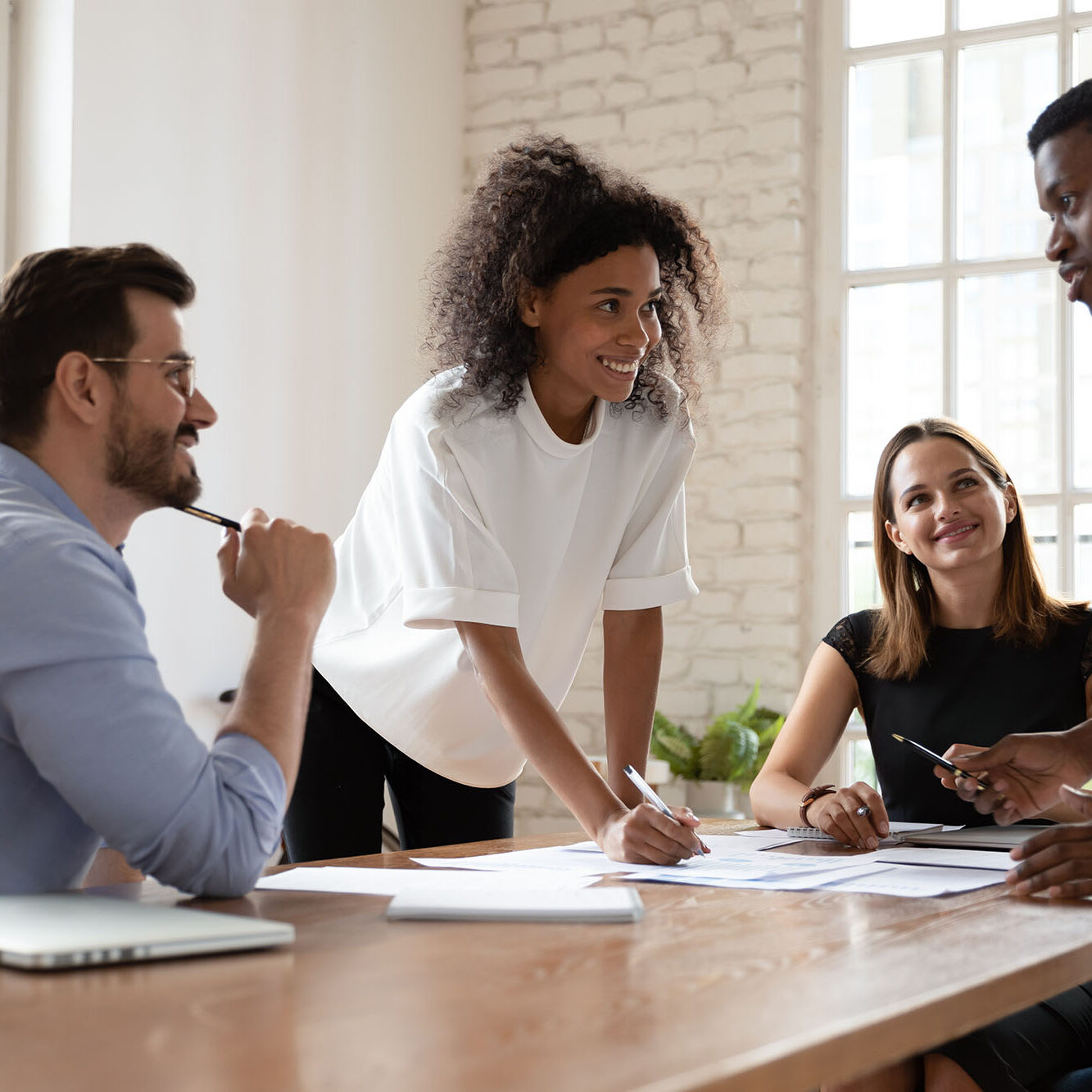Friendly mixed race company workers listening to smiling african american young employee, sharing project ideas at brainstorming meeting. Skilled biracial professional proposing good problem solution.