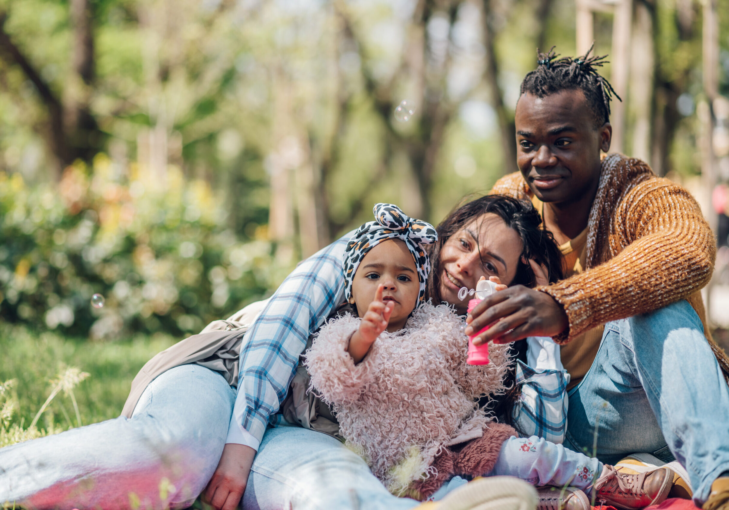 Beautiful baby girl enjoying making soap bubbles in the park while spending a beautiful sunny day with her diverse parents outside. Happy multiracial family concept.