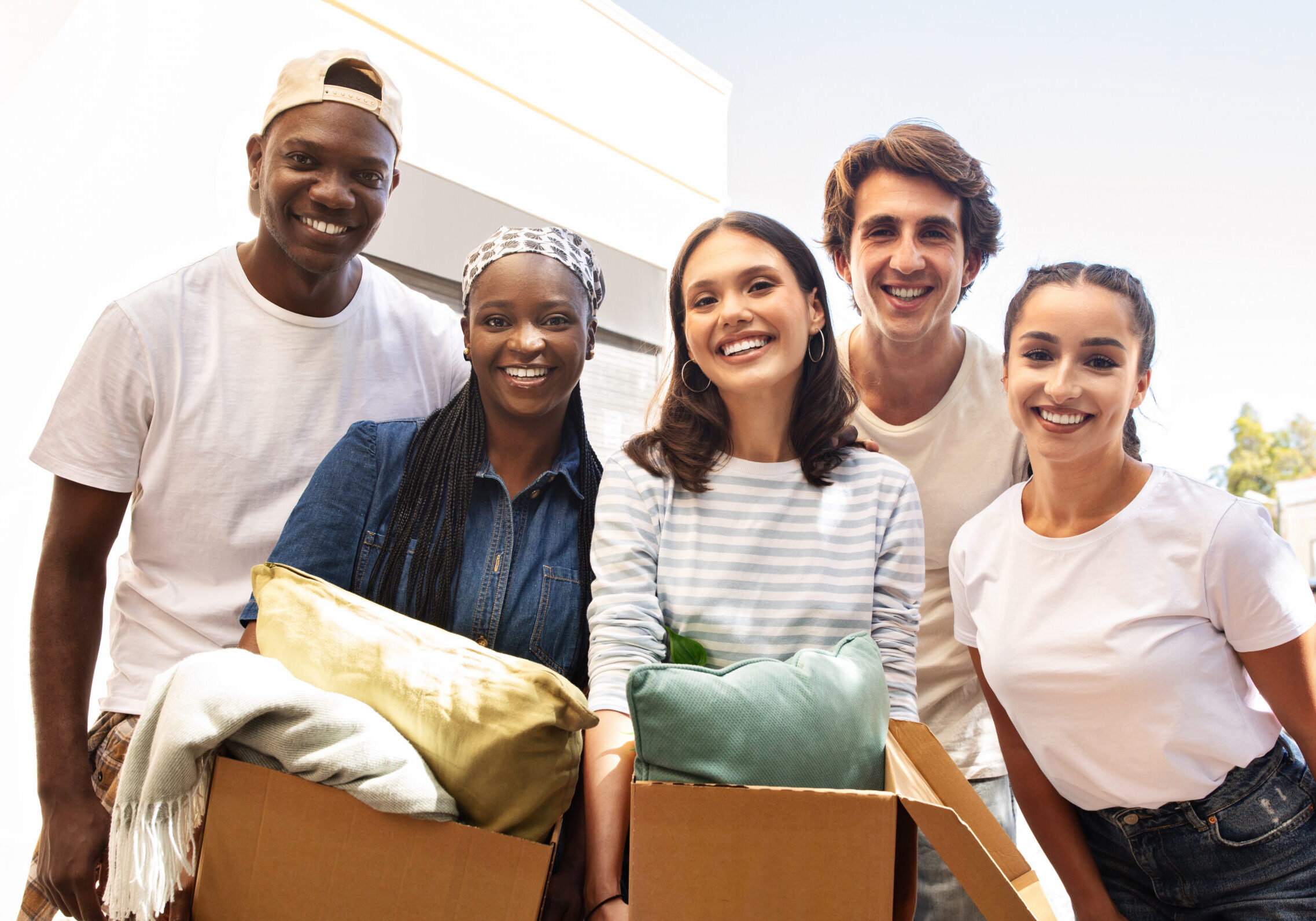 Cheerful multicultural group of students two young guys and three ladies holding boxes with belongings, standing by building, smiling at camera, friends hellp with moving to new apartment, relocation